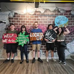 Escape room victory group photo of five people standing against a graffiti-covered brick wall holding colorful hand-painted signs reading WELP... WE tried, They should've listened to ME!, LOCKED UP, Victory, and Guess what? WE WON!