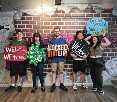 Escape room victory group photo of five people standing against a graffiti-covered brick wall holding colorful hand-painted signs reading WELP... WE tried, They should've listened to ME!, LOCKED UP, Victory, and Guess what? WE WON!