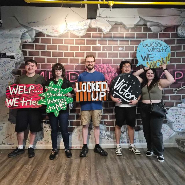 Escape room victory group photo of five people standing against a graffiti-covered brick wall holding colorful hand-painted signs reading WELP... WE tried, They should've listened to ME!, LOCKED UP, Victory, and Guess what? WE WON!