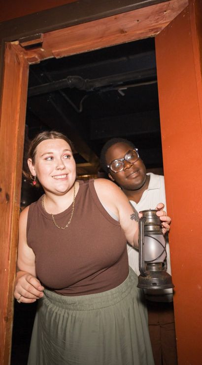Two people playfully peeking through a rustic wooden doorway in a warm-lit room, one holding a vintage lantern with shelves and textured wall panel visible.