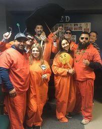 Group of eight adults in orange jumpsuit costumes at an indoor office Halloween party, posing and smiling with emoji props, sunglasses, beads and an umbrella for a playful photo