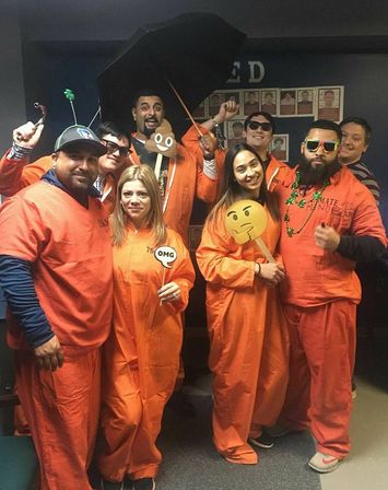 Group of eight adults in orange jumpsuit costumes at an indoor office Halloween party, posing and smiling with emoji props, sunglasses, beads and an umbrella for a playful photo