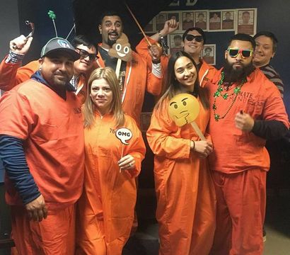 Group of eight adults in orange jumpsuit costumes at an indoor office Halloween party, posing and smiling with emoji props, sunglasses, beads and an umbrella for a playful photo