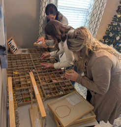 Three people choosing charms and beads from wooden compartment trays at a cozy indoor bracelet-making station beside a decorated Christmas tree.