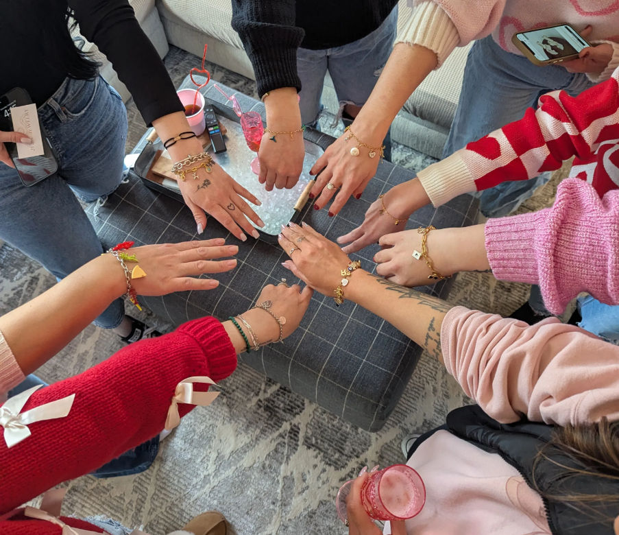Top-down view of a friends' gathering in a cozy living room — hands arranged in a circle over a gray plaid ottoman showcasing charm bracelets, rings and tattoos, with pink drinks and a smartphone nearby.