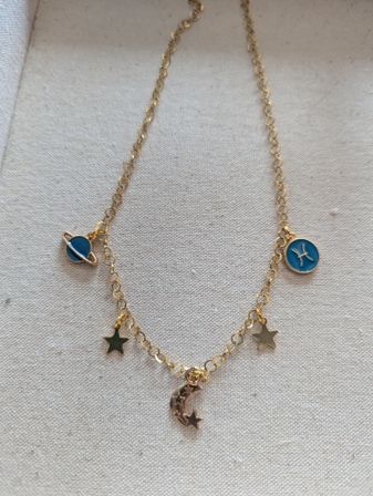 Delicate gold charm necklace with crescent moon, tiny star charms and blue enamel planet and Pisces discs on beige fabric background
