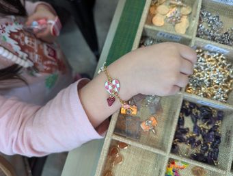 Child's wrist wearing a gold charm bracelet with a pink-checkered heart and red strawberry charm, reaching into a burlap divided jewelry tray filled with metal and gemstone charms on a market-style display.