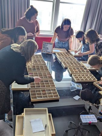 Group of women gathered around a high-rise hotel table with a city view selecting beads and charms from wooden compartment trays during a DIY charm-bar bracelet workshop.