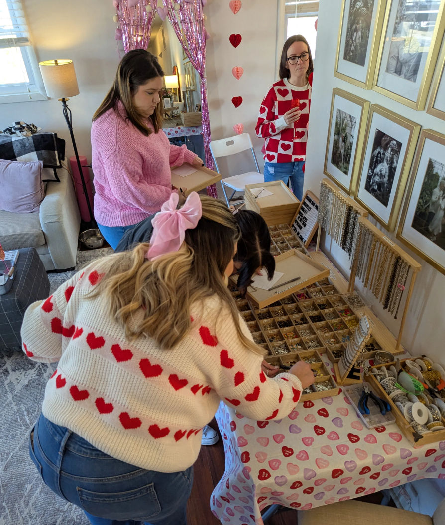 Women browsing handmade jewelry trays at a cozy Valentine’s-themed pop-up in a living room, wearing heart-pattern sweaters and pink decor.