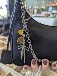 Black mini shoulder bag with chunky silver chain and dangling charms — yellow smiley face, round brass charm with an 'm' and a silver bow — held by a hand with glittery mauve nails in a boutique display setting.