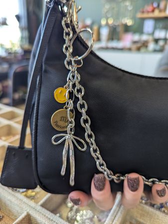 Black mini shoulder bag with chunky silver chain and dangling charms — yellow smiley face, round brass charm with an 'm' and a silver bow — held by a hand with glittery mauve nails in a boutique display setting.