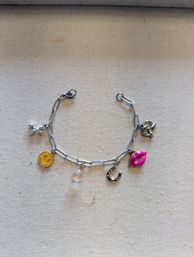 Flat-lay close-up of a silver charm bracelet on beige fabric, featuring a yellow smiley face charm, small round pearl, horseshoe, bright pink lips and a tiny airplane charm.