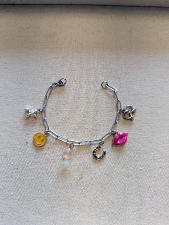 Flat-lay close-up of a silver charm bracelet on beige fabric, featuring a yellow smiley face charm, small round pearl, horseshoe, bright pink lips and a tiny airplane charm.