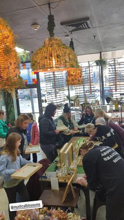 Group of people at a busy indoor craft workshop assembling jewelry at display tables in a bright, plant-filled storefront with hanging woven lamps and large windows.