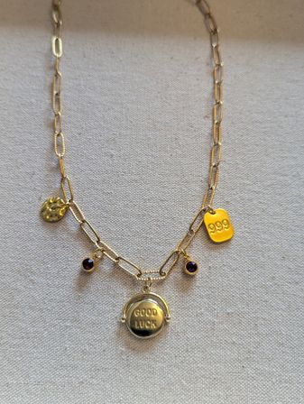 Gold paperclip-chain necklace displayed on beige linen, featuring a round pendant stamped "GOOD LUCK", a small hammered coin charm, a rectangular tag stamped "999" and two tiny purple gemstone drops.