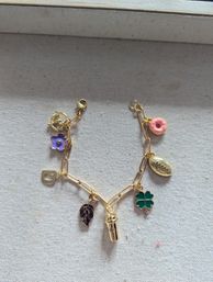 Close-up flatlay of a gold charm bracelet on beige textured canvas, playful jewelry accessory with colorful charms: purple flower, pink donut, green four-leaf clover, dark leaf, mini drink tumbler and a small gold oval tag.