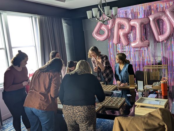 Group crafting at a bridal shower jewelry-making workshop in a hotel room — people gathered around trays of beads and tools, pink 'BRIDE' balloons and iridescent fringe backdrop