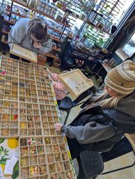 Overhead view in a cozy craft shop: two shoppers and a child at a table of compartment trays packed with colorful beads, charms, and jewelry findings for DIY jewelry making.