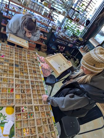 Overhead view in a cozy craft shop: two shoppers and a child at a table of compartment trays packed with colorful beads, charms, and jewelry findings for DIY jewelry making.