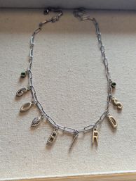 Silver paperclip-chain charm necklace laid flat on beige linen, with dangling letter charms (B, I, R, D), a small football charm and two green gemstone accents — personalized jewelry flatlay.