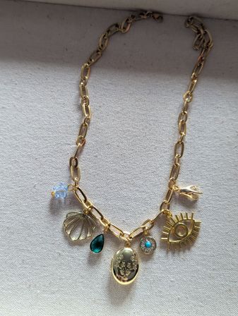 Gold charm necklace flatlay on beige linen background, chunky oval-link chain with dangling shell charm, green teardrop gem, engraved floral locket, small turquoise disc, sun-eye charm and tiny blue crystal bead.