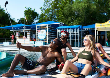 Three friends in swimwear taking a selfie while lounging on paddleboards and kayaks at a sunny lakeside dock, sunglasses on and watercraft racks and blue canopies in the background — summer waterfront recreation.