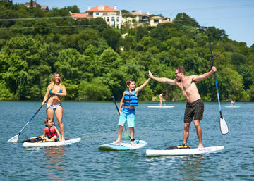 Sunny lake scene with three people stand-up paddleboarding near a tree-covered shoreline and hillside homes — man high-fiving a child while a woman and toddler share a nearby board.