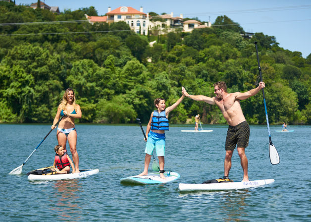 Sunny lake scene with three people stand-up paddleboarding near a tree-covered shoreline and hillside homes — man high-fiving a child while a woman and toddler share a nearby board.