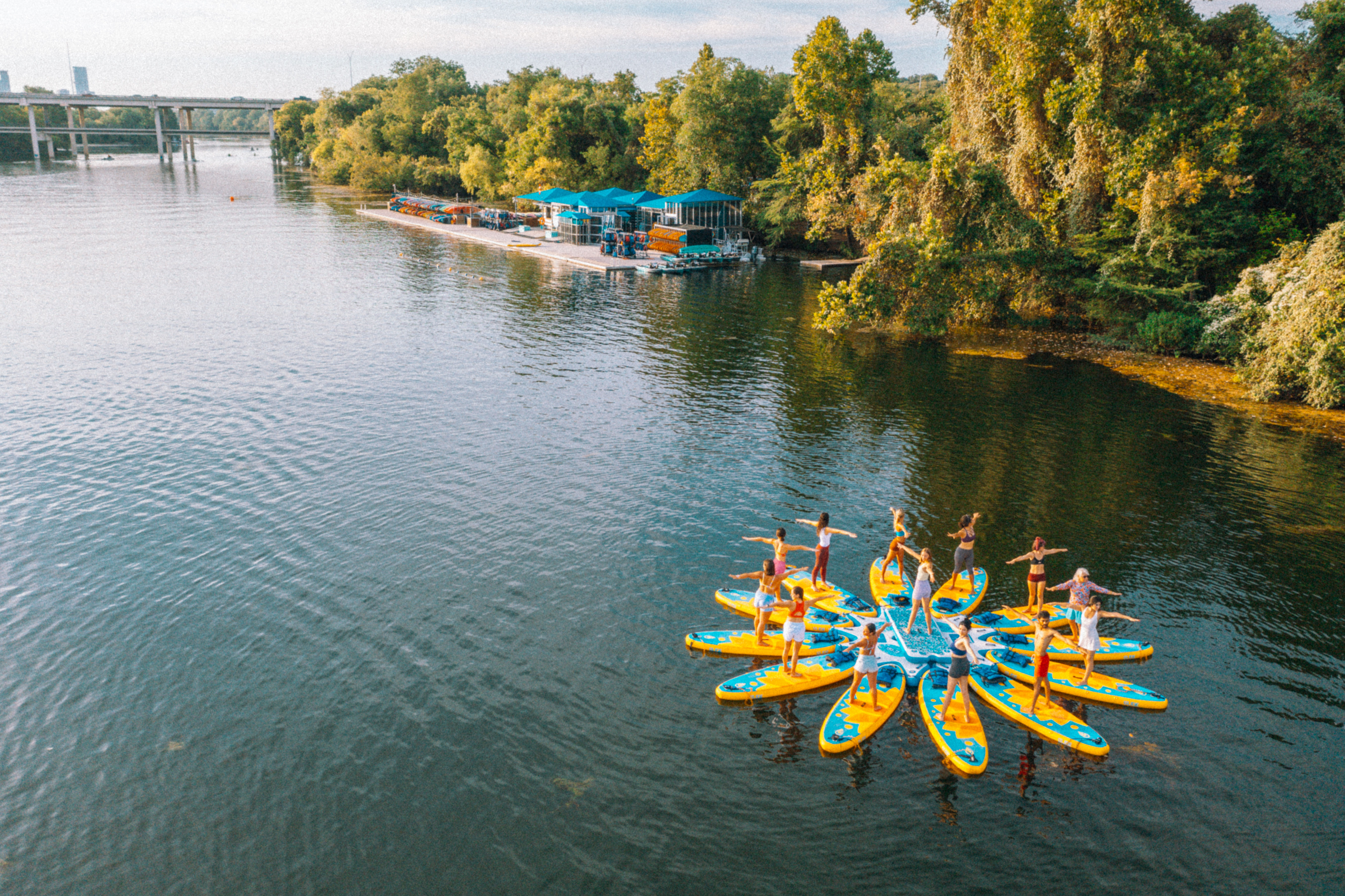 Aerial view of a group doing stand-up paddleboard yoga on bright yellow-and-blue boards arranged like a flower on a calm river near a tree-lined shore and dock.