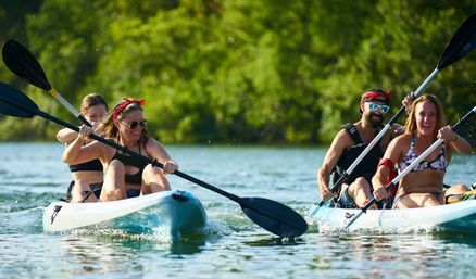 Four friends paddling two tandem kayaks on a sunny lake, smiling and splashing near a green, tree-lined shore — summer outdoor kayaking adventure.