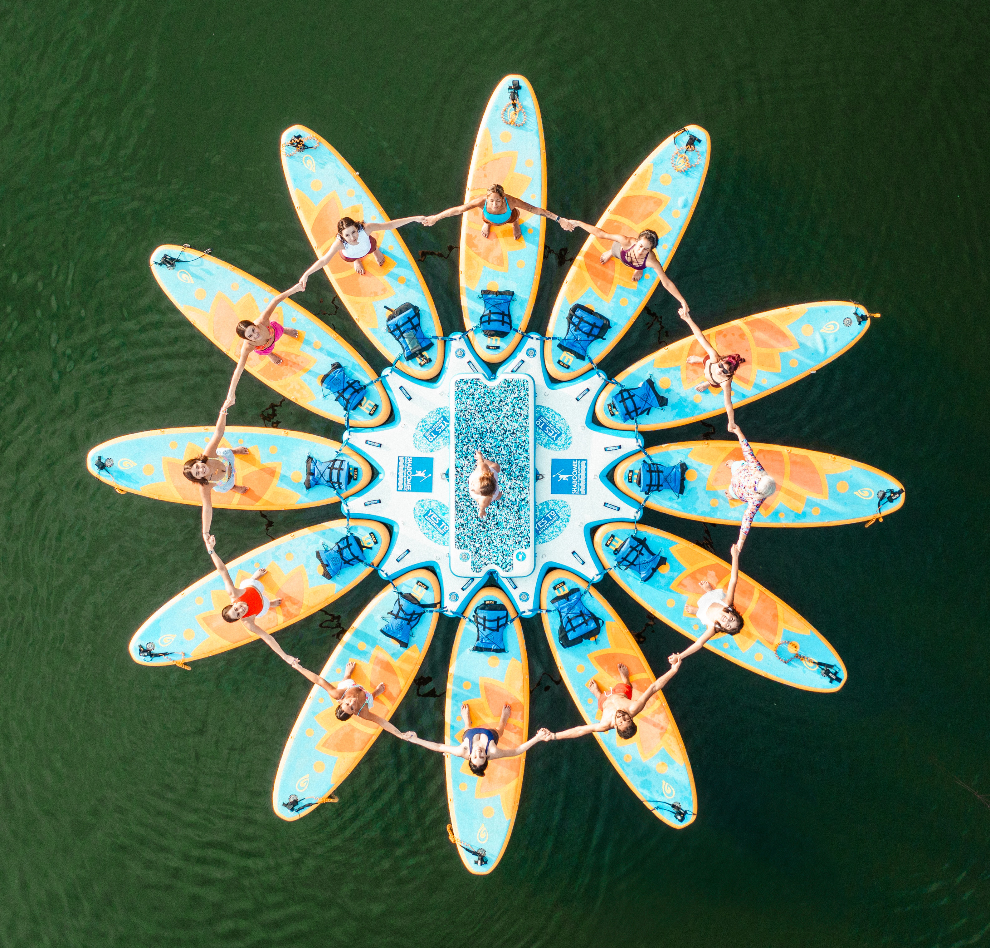 Aerial drone view of a sunflower-shaped ring of colorful stand-up paddleboards on a green lake, people holding hands in a circular formation