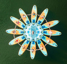 Aerial drone view of a sunflower-shaped ring of colorful stand-up paddleboards on a green lake, people holding hands in a circular formation