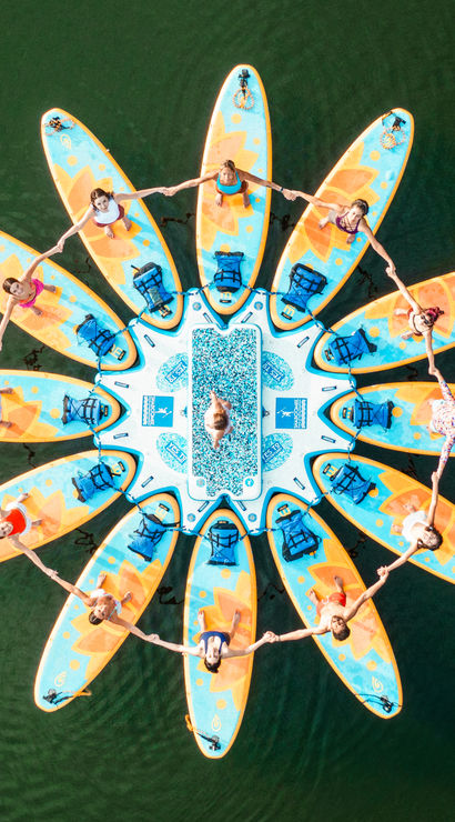 Aerial drone view of a sunflower-shaped ring of colorful stand-up paddleboards on a green lake, people holding hands in a circular formation