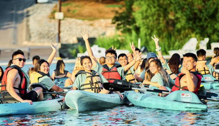Group of smiling young adults kayaking on a sunny lake, wearing life jackets and waving from turquoise recreational kayaks—summer lakeside outdoor activity.