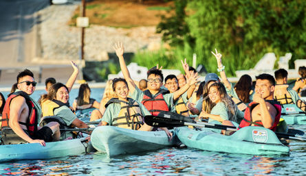 Group of smiling young adults kayaking on a sunny lake, wearing life jackets and waving from turquoise recreational kayaks—summer lakeside outdoor activity.