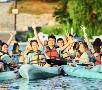 Group of smiling young adults kayaking on a sunny lake, wearing life jackets and waving from turquoise recreational kayaks—summer lakeside outdoor activity.