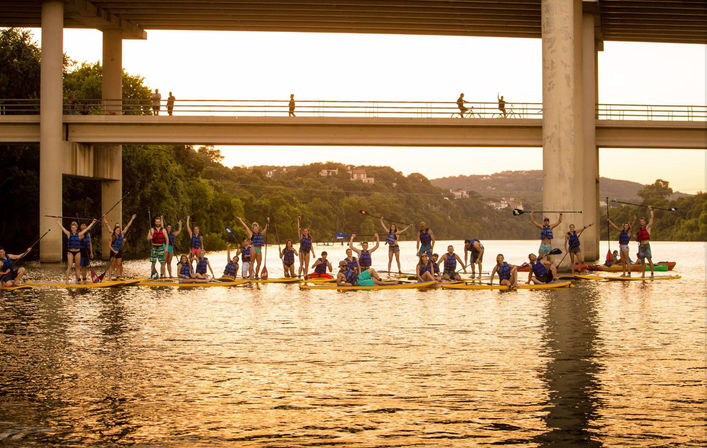 Large group of paddleboarders in life jackets on a calm river at golden sunset beneath a concrete bridge, cheering and holding paddles with tree-covered hills in the background.