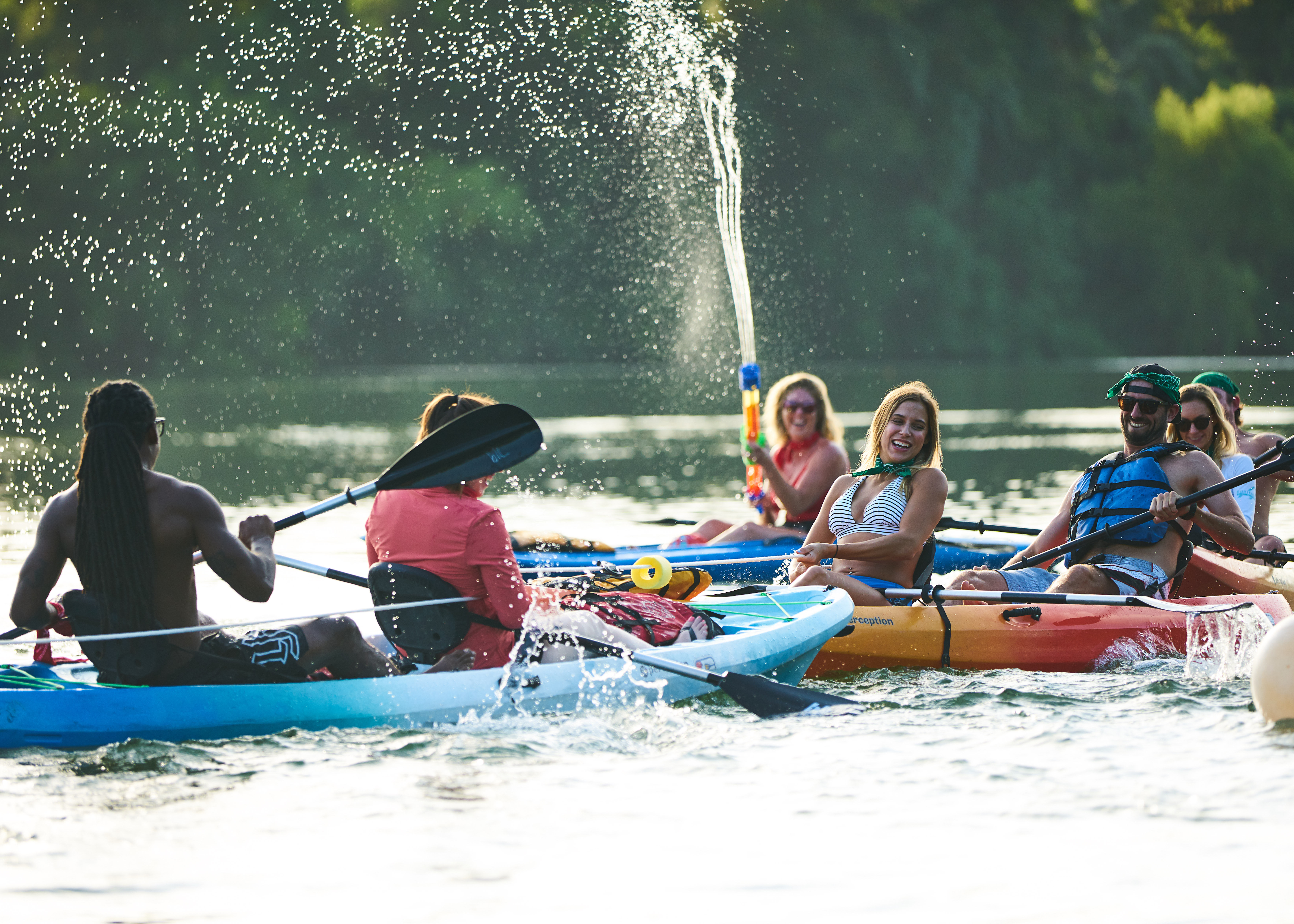 Smiling kayakers paddling and splashing on a sunlit lake in colorful kayaks, one person firing a water gun high into the air