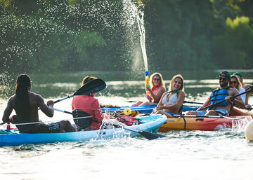 Smiling kayakers paddling and splashing on a sunlit lake in colorful kayaks, one person firing a water gun high into the air