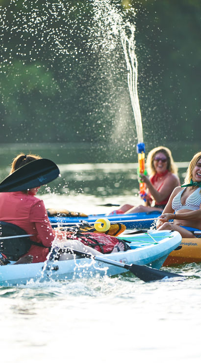 Smiling kayakers paddling and splashing on a sunlit lake in colorful kayaks, one person firing a water gun high into the air