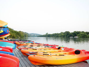 Colorful kayaks and paddles lined up on a wooden dock at a calm lake, ready to launch against a green tree-lined shoreline and distant hills