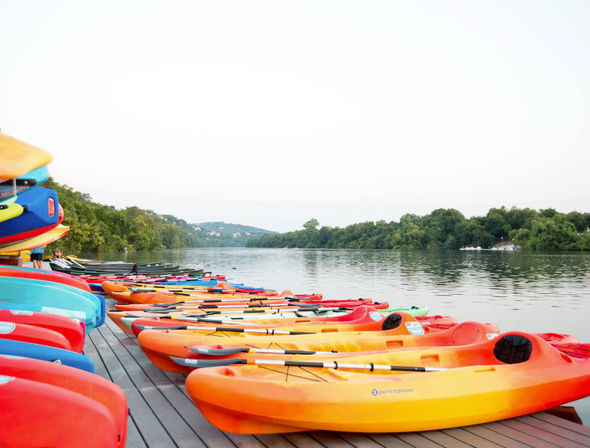Colorful kayaks and paddles lined up on a wooden dock at a calm lake, ready to launch against a green tree-lined shoreline and distant hills