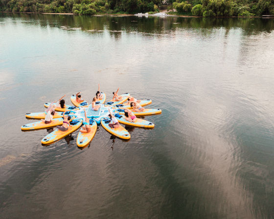 Group doing paddleboard yoga on yellow stand-up paddleboards arranged in a sunflower circle on a calm lake with a tree-lined shoreline