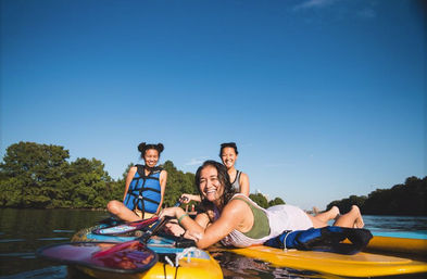 Three friends paddleboarding on a calm river on a sunny summer day, two wearing blue life jackets and one lying on a yellow board, smiling with a tree-lined shore and clear blue sky