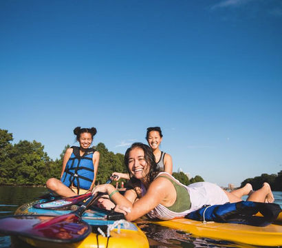Three friends paddleboarding on a calm river on a sunny summer day, two wearing blue life jackets and one lying on a yellow board, smiling with a tree-lined shore and clear blue sky