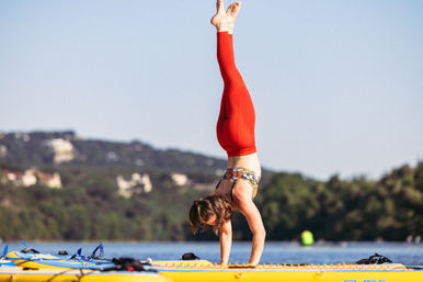 Woman in red leggings doing a handstand on a yellow paddleboard on a calm lake with tree-lined hills in the background