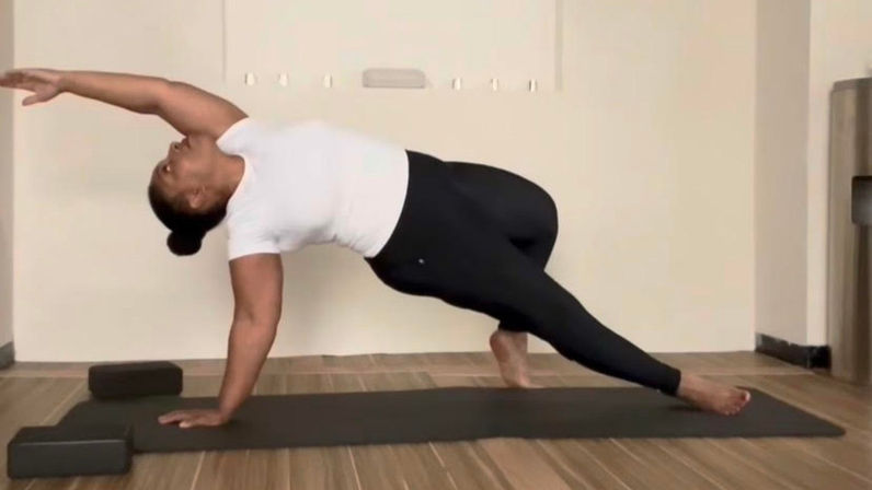 Person performing a dynamic side-plank yoga pose on a mat in an indoor workout space, reaching top arm overhead for core strength and balance