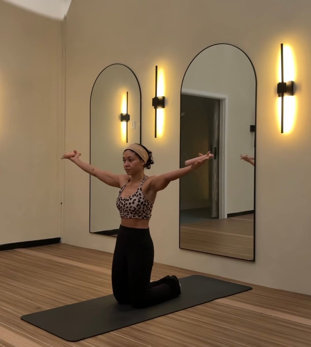 Person kneeling on a black mat in a bright indoor yoga studio, arms outstretched in a stretch, leopard-print sports bra and black leggings, arched wall mirrors and modern vertical sconces over wood floor