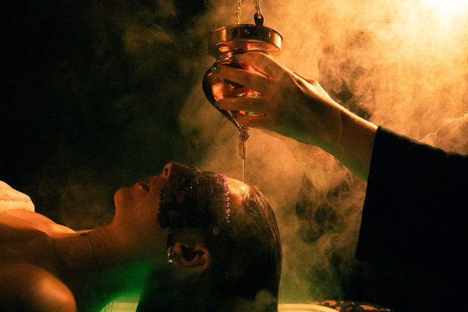 Reclined person receiving warm oil drip from a brass vessel in an amber-lit, smoky spa head treatment for deep relaxation.