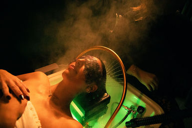 Person reclining in a salon sink for a steaming scalp treatment under a circular shower arm, green light and billowing steam creating a moody spa atmosphere.